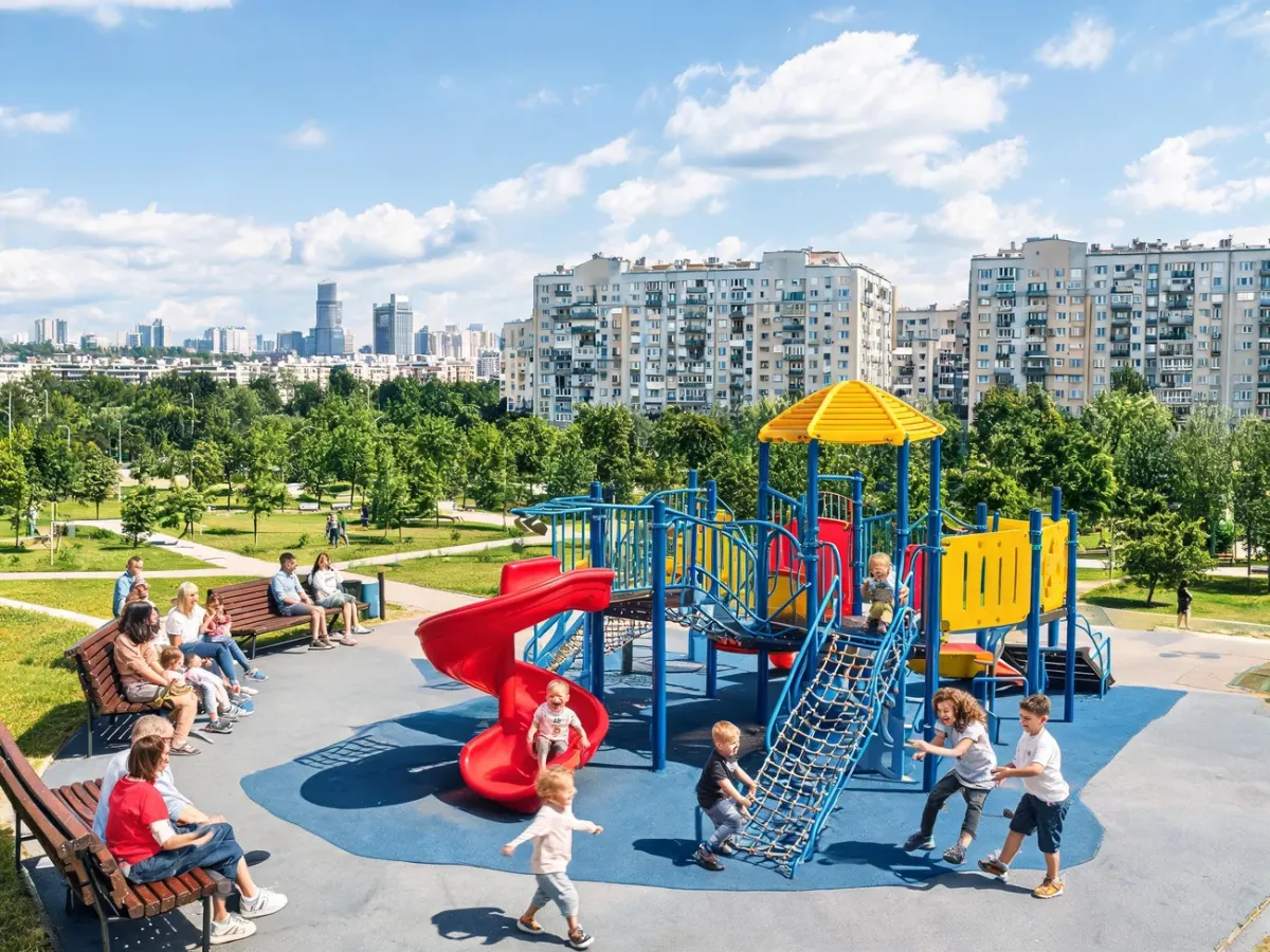 Dzieci bawiące się na kolorowym placu zabaw w polskim mieście, w tle park, bloki i słoneczne, rodzinne otoczenie.